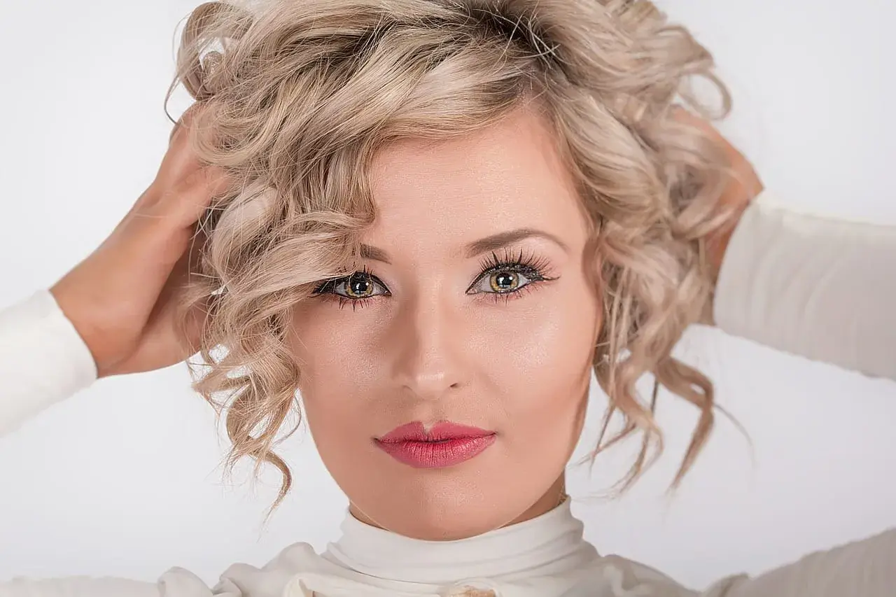 Kobieta z kręconymi blond włosami, jej spojrzenie jest nonszalanckie, a dłonie unoszą się ku głowie.