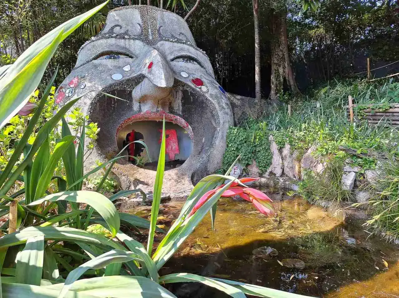 Scultura di una testa gigante con bocca spalancata nel giardino botanico Gardone Riviera, con piante rigogliose e uno stagno.