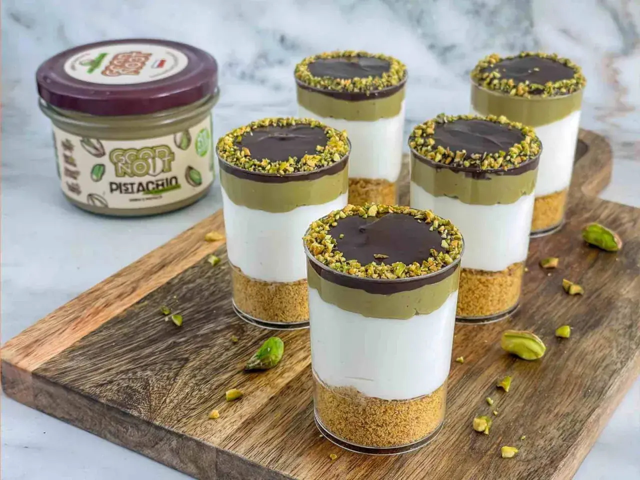 Deser pistacjowy w szklankach z polewą czekoladową i posypką z pistacji. Słoik z kremem pistacjowym w tle.