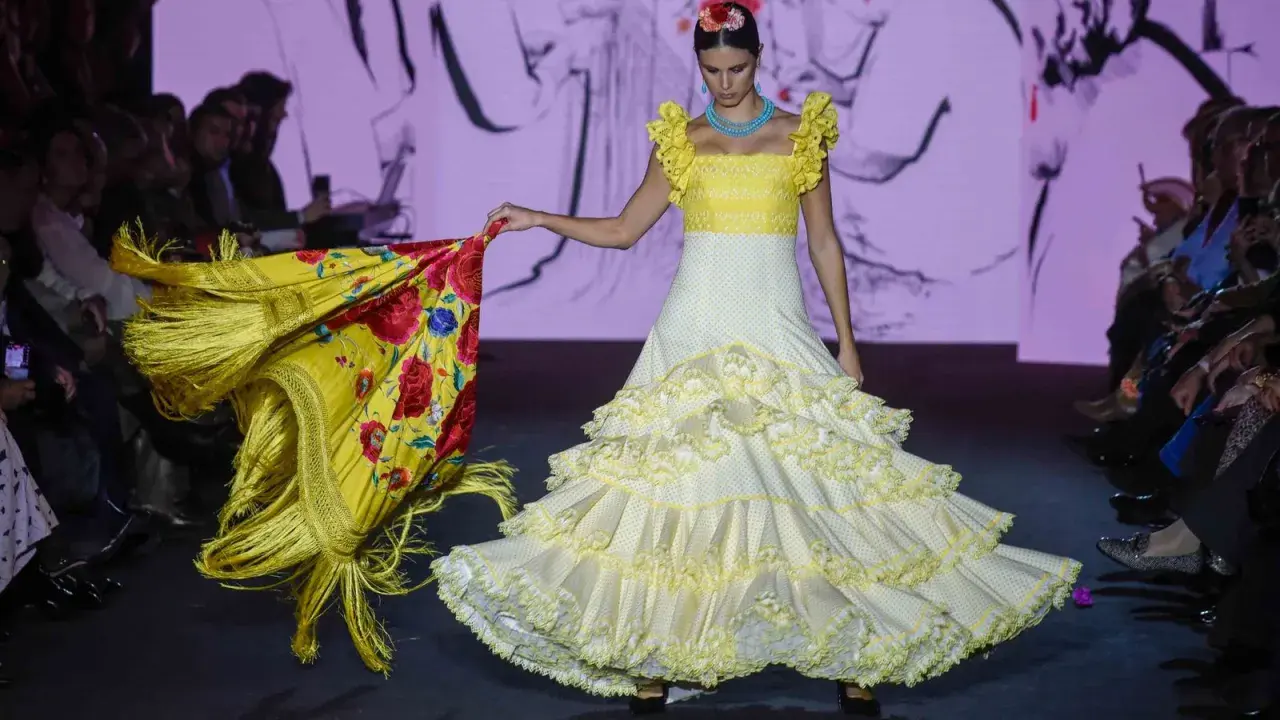Modelo desfila en la pasarela We Love Flamenco con un vestido blanco de lunares amarillos y volantes, portando un mantón amarillo con flores rojas.