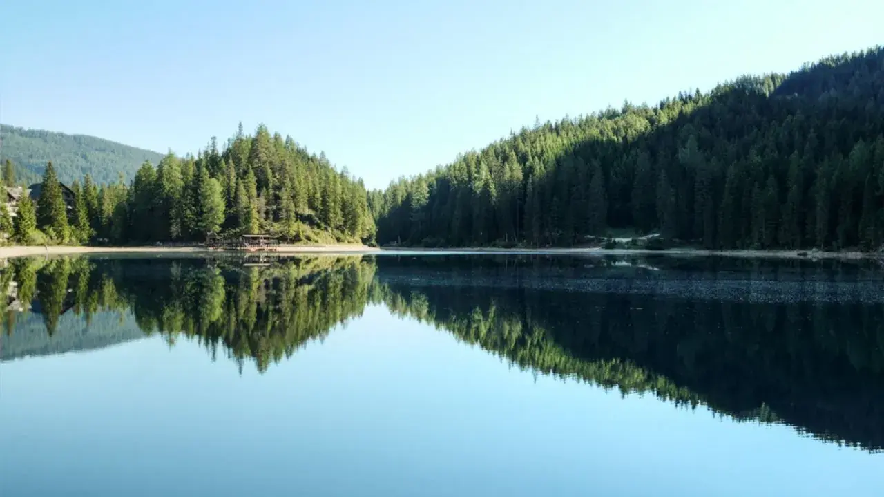 Spokojna tafla jeziora odbija gęsty las i góry. Widok na malowniczą kotlinę z drewnianym pomostem.