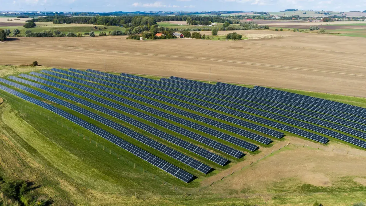 En stor solcellspark på åkermark, med rader av solpaneler som sträcker sig över grönskande och brun jordbruksmark.