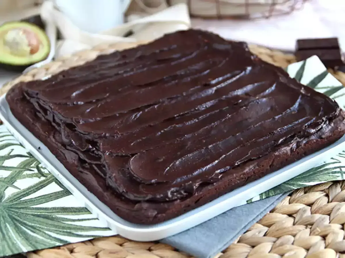 Puszysty brownie z awokado, polane błyszczącą polewą czekoladową. Obok połówka awokado i kawałek gorzkiej czekolady.