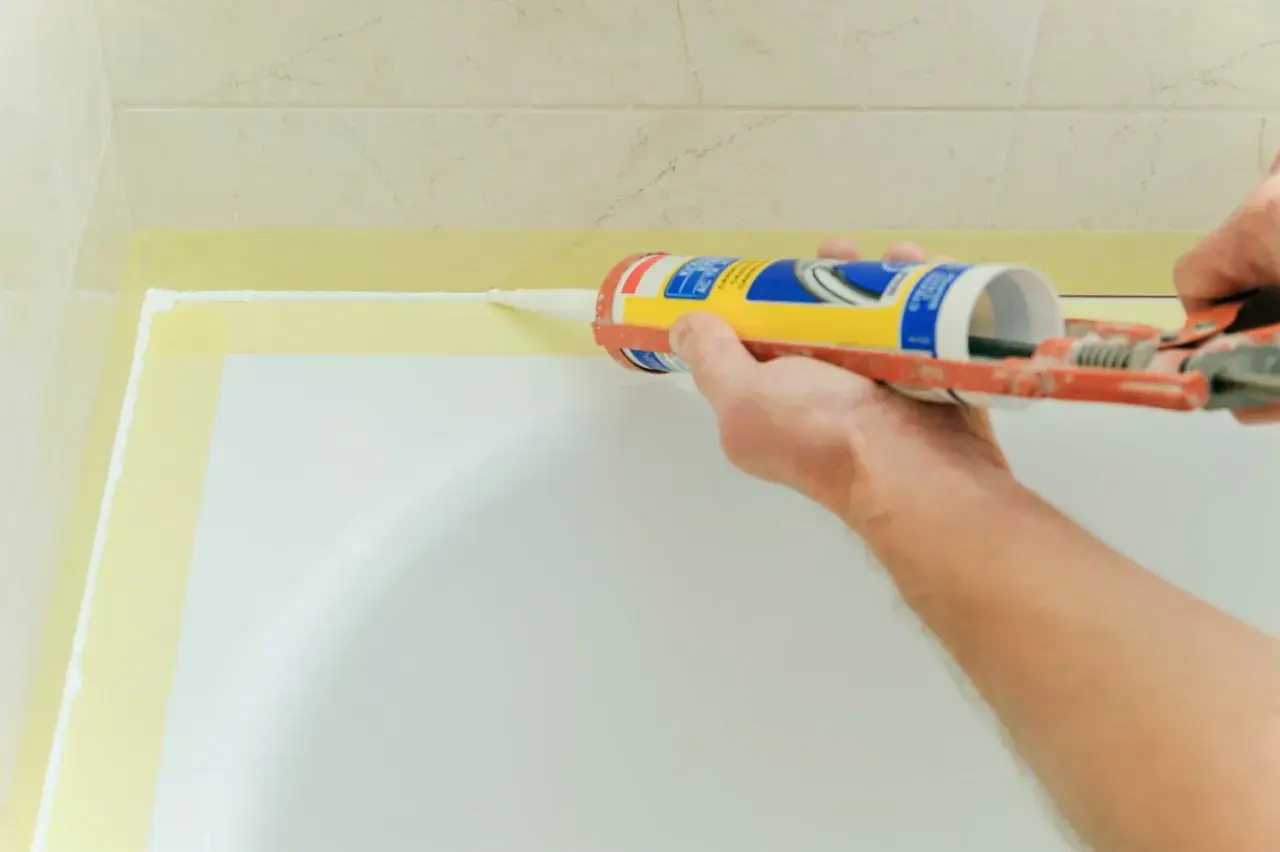 Aplikacja silikonu do brodzika. Ręka trzymająca pistolet z silikonem, nakładająca białą masę na krawędź brodzika.