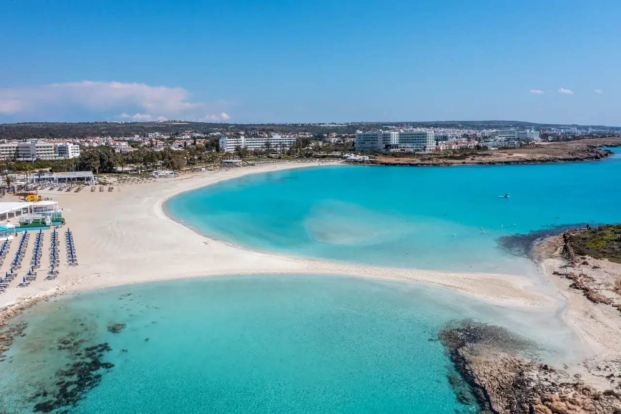 Piękna plaża Ayia Napa z turkusową wodą i białym piaskiem, otoczona hotelami i zielenią.