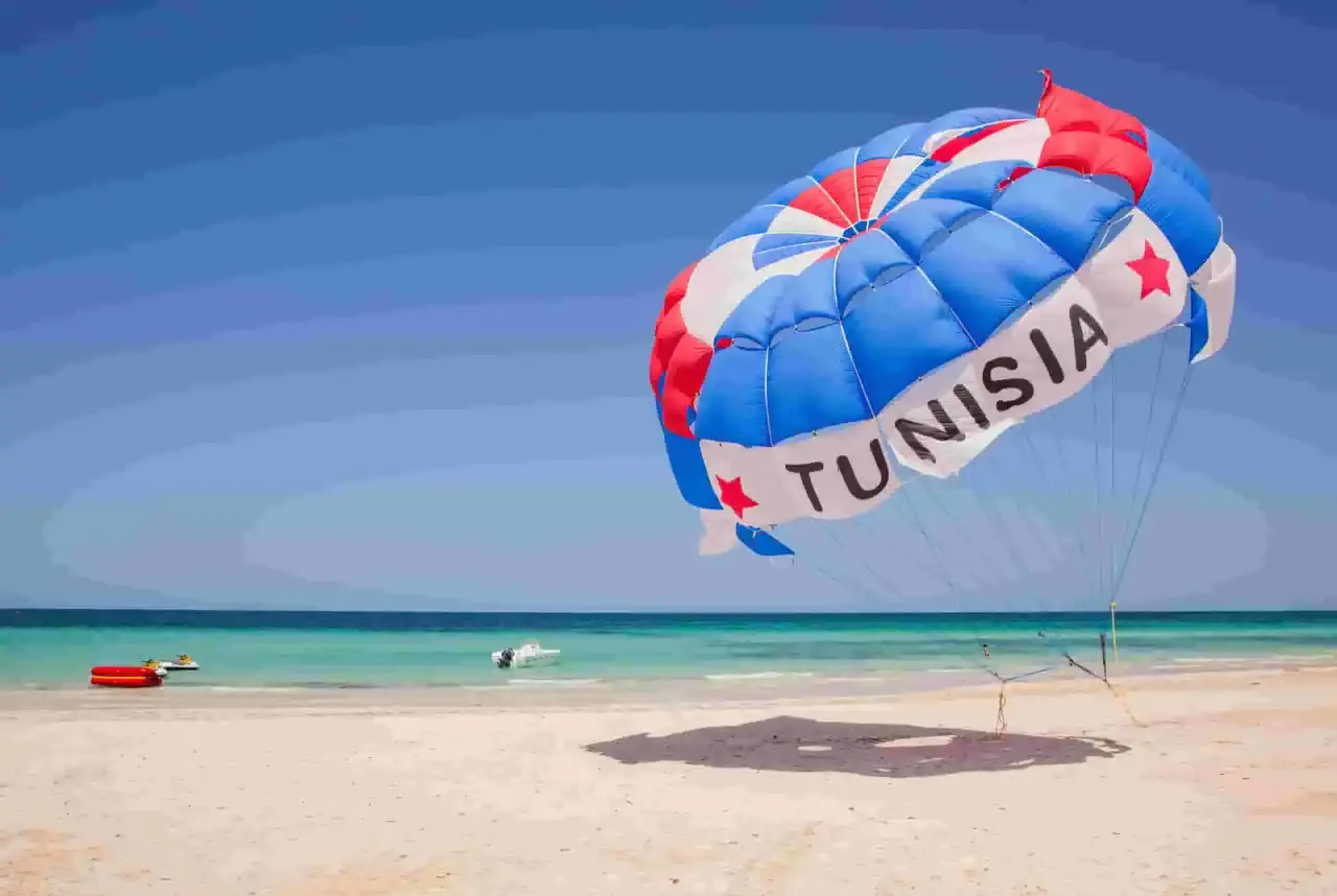 Kolorowy spadochron z napisem "TUNISIA" unosi się nad plażą, zapowiadając wspaniałe dżerba loty. W tle turkusowe morze i piaszczysta plaża.