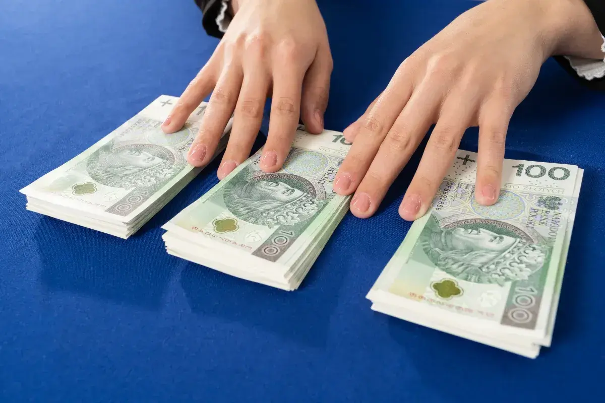 Dłonie nad stosem banknotów 100 zł. Symbolizuje to darowiznę między małżonkami, wsparcie finansowe lub oszczędności.