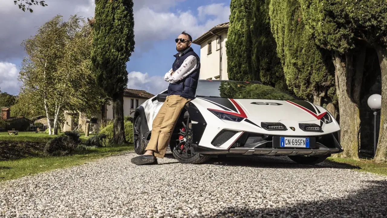 Mężczyzna opiera się o białe Lamborghini Sterrato z czerwono-czarnymi akcentami, zaparkowane na żwirowej drodze wśród drzew.