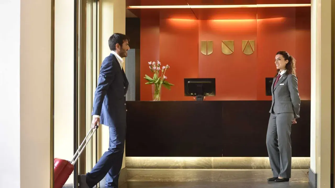 Uomo con valigia rossa alla reception, pronto per l'early check-in. La receptionist sorride accogliendolo.