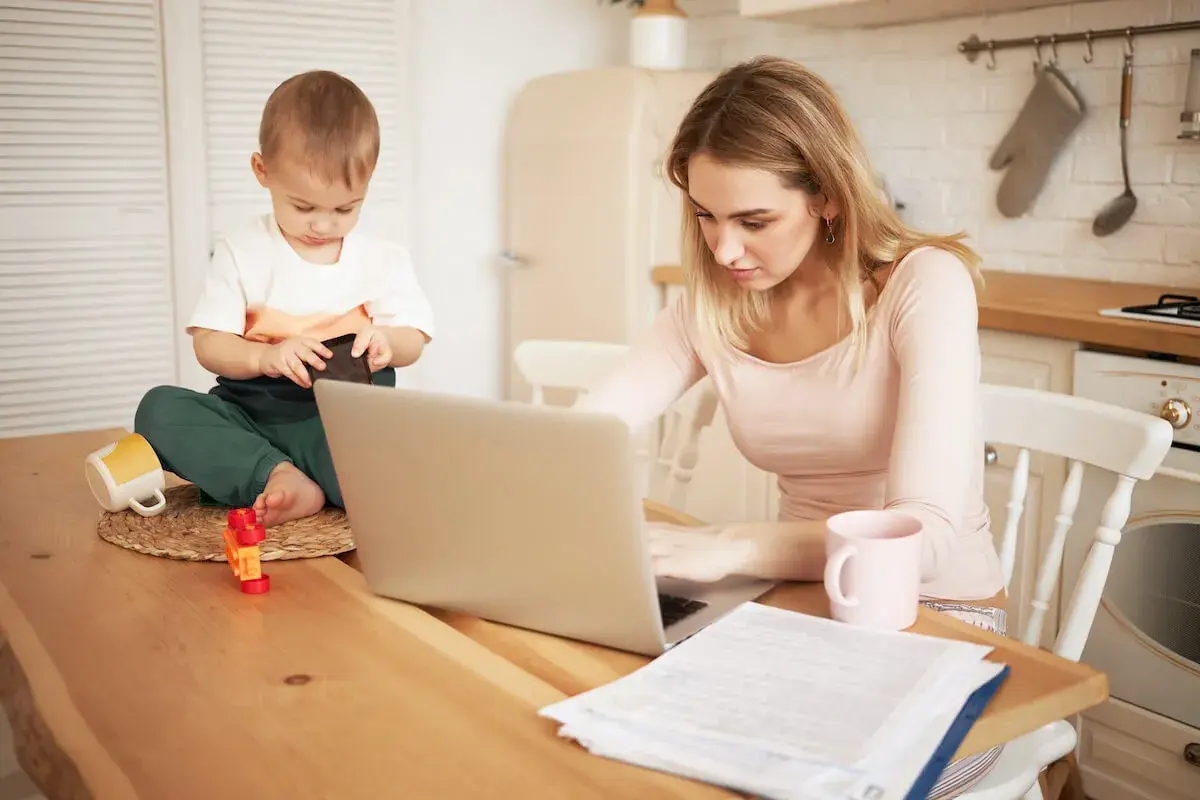 Młoda mama pracuje na laptopie, podczas gdy jej małe dziecko bawi się obok. Czy na urlopie macierzyńskim można pracować? Tak, często jest to możliwe.