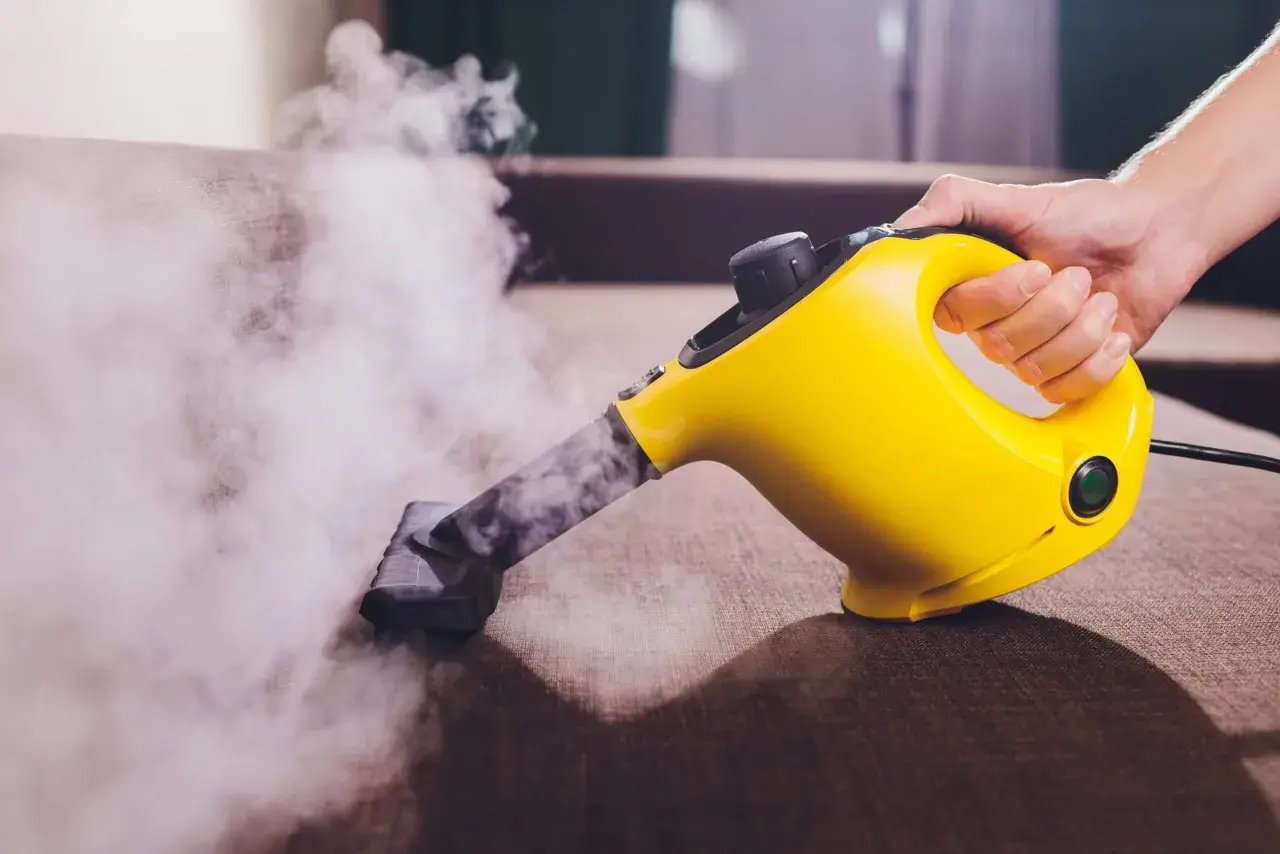 Mano sujetando una limpiadora a vapor amarilla, desprendiendo vapor sobre un sofá marrón.