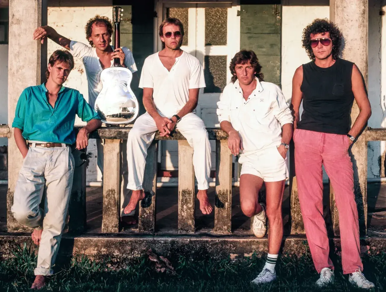 Członkowie Dire Straits pozują na tle budynku. Jeden z nich trzyma gitarę.