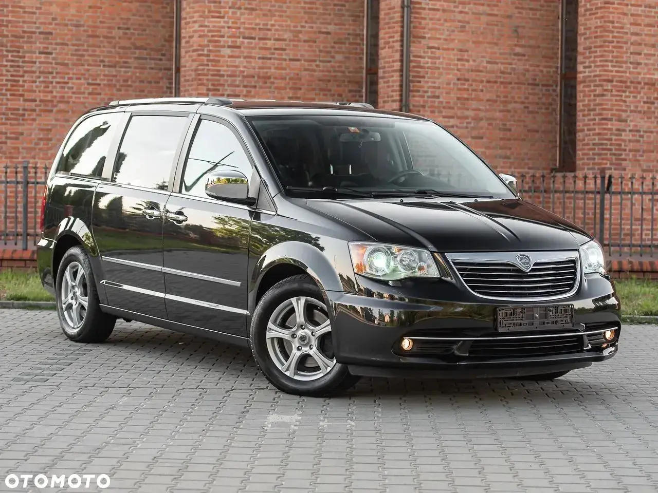 Czarny Lancia Voyager stoi na brukowanym podjeździe przed ceglanym budynkiem.