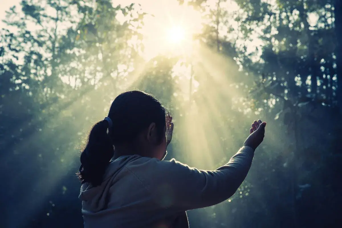 Una niña con coleta, con las manos alzadas, parece soñar con espíritus mientras la luz del sol atraviesa los árboles.
