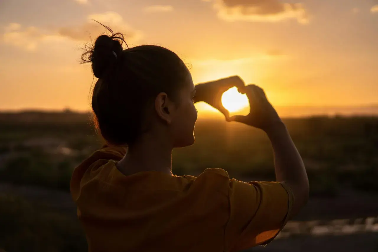 Silueta de mujer haciendo un corazón con las manos al atardecer, reflejando su actitud positiva ante la vida.