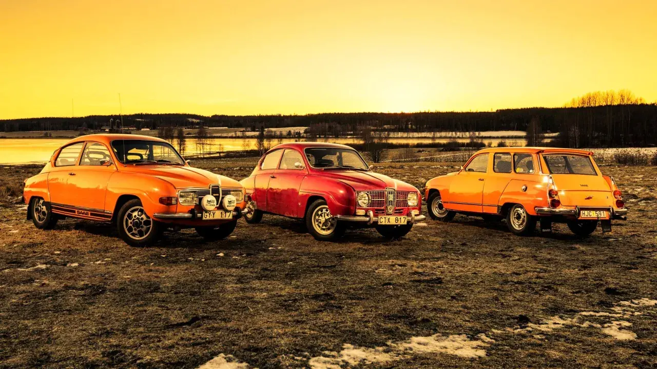 Tre klassiska Saab-bilar, en röd och två orange, står uppradade i solnedgången. En laban bil som väcker nostalgi.