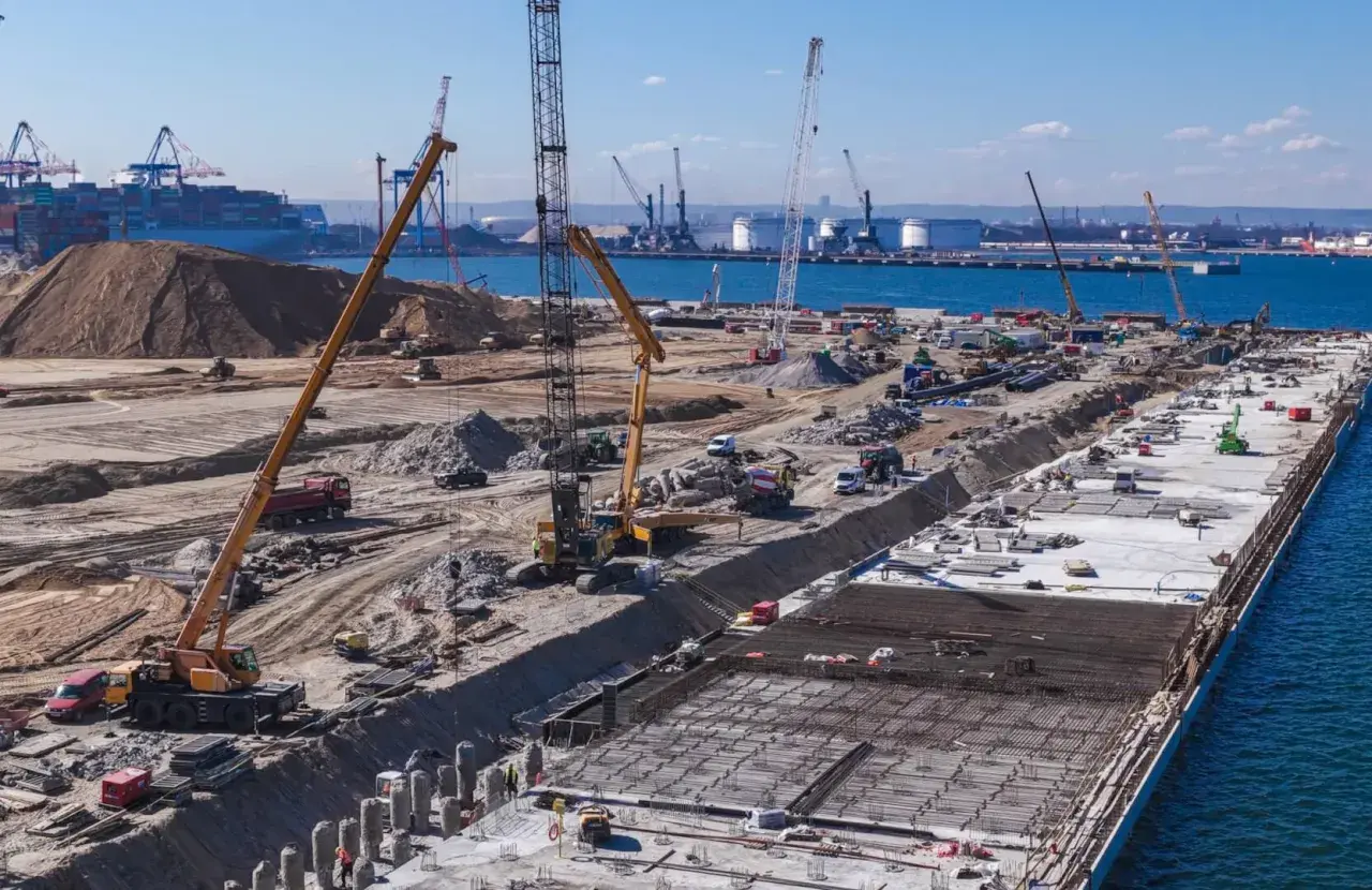 Budowa dużego portu w Japonii. Dźwigi, koparki i ciężarówki pracują na placu budowy, tworząc nową infrastrukturę.