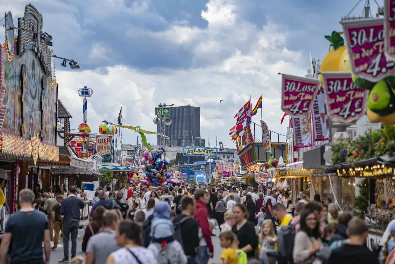 Buntes Volksfest mit vielen Menschen, Fahrgeschäften und Buden. Ein Hamburger-Stand ist nicht zu sehen, aber die Stimmung ist ausgelassen.