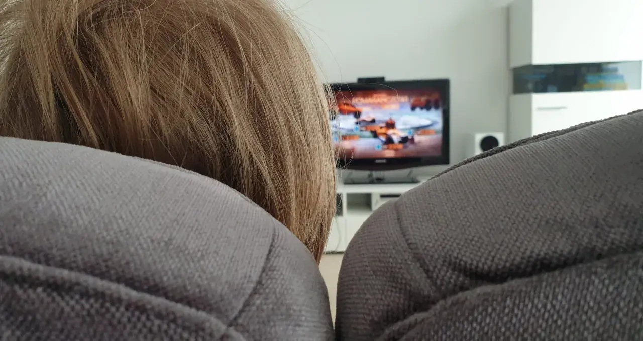 Niemowlę z blond włosami siedzi na kanapie, a w tle widać włączony telewizor z grą.