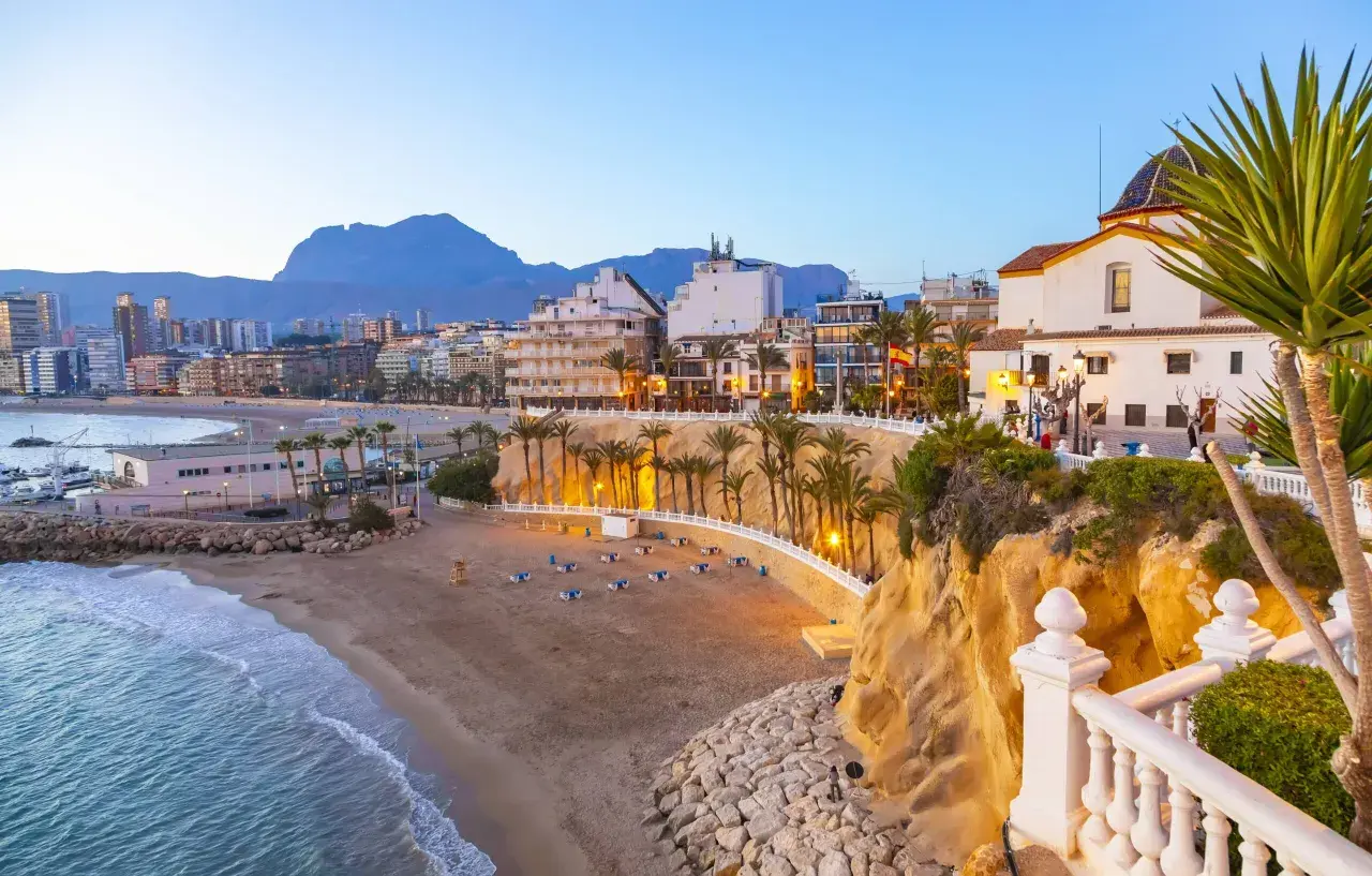 Atardecer en la playa Poniente de Benidorm, con el mar azul, la arena dorada, palmeras y edificios modernos.
