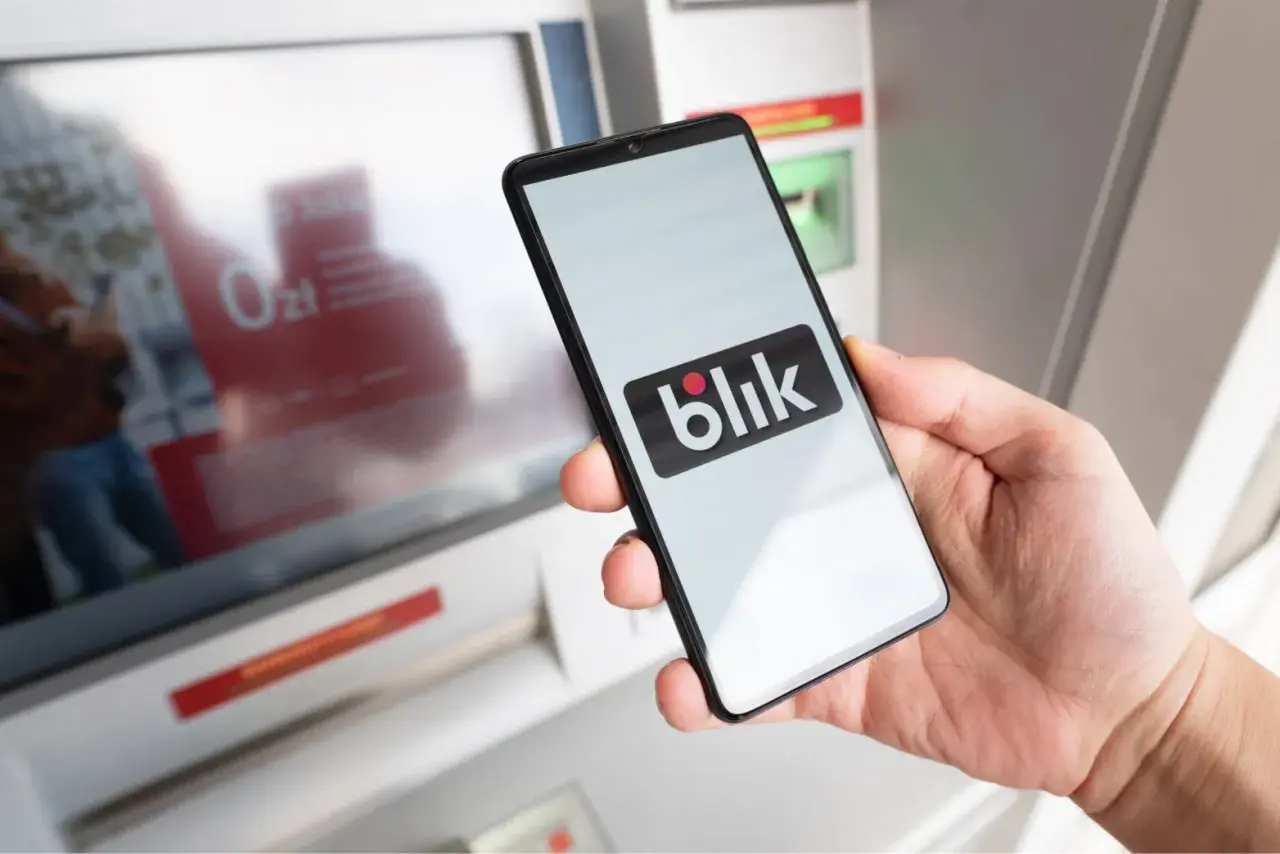 Dłoń trzyma smartfon z logo Blik. Czy Blik jest tylko w Polsce? Płatność zbliżeniowa przy bankomacie.