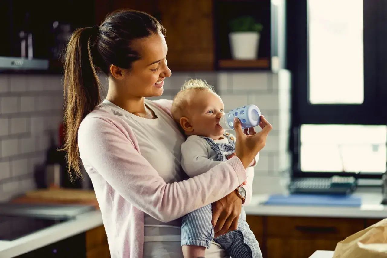 Uśmiechnięta mama karmi butelką niemowlę. Kiedy można podać koperek dla noworodka? To pytanie nurtuje wielu rodziców.