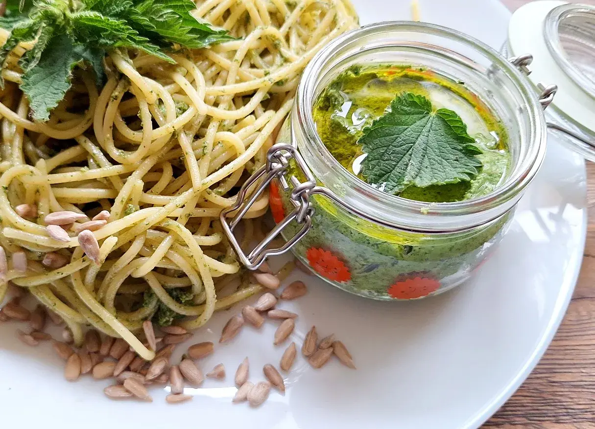 Makaron z domowym pesto z pokrzywy, ozdobiony świeżymi liśćmi i pestkami słonecznika. Pesto w słoiczku wygląda apetycznie.