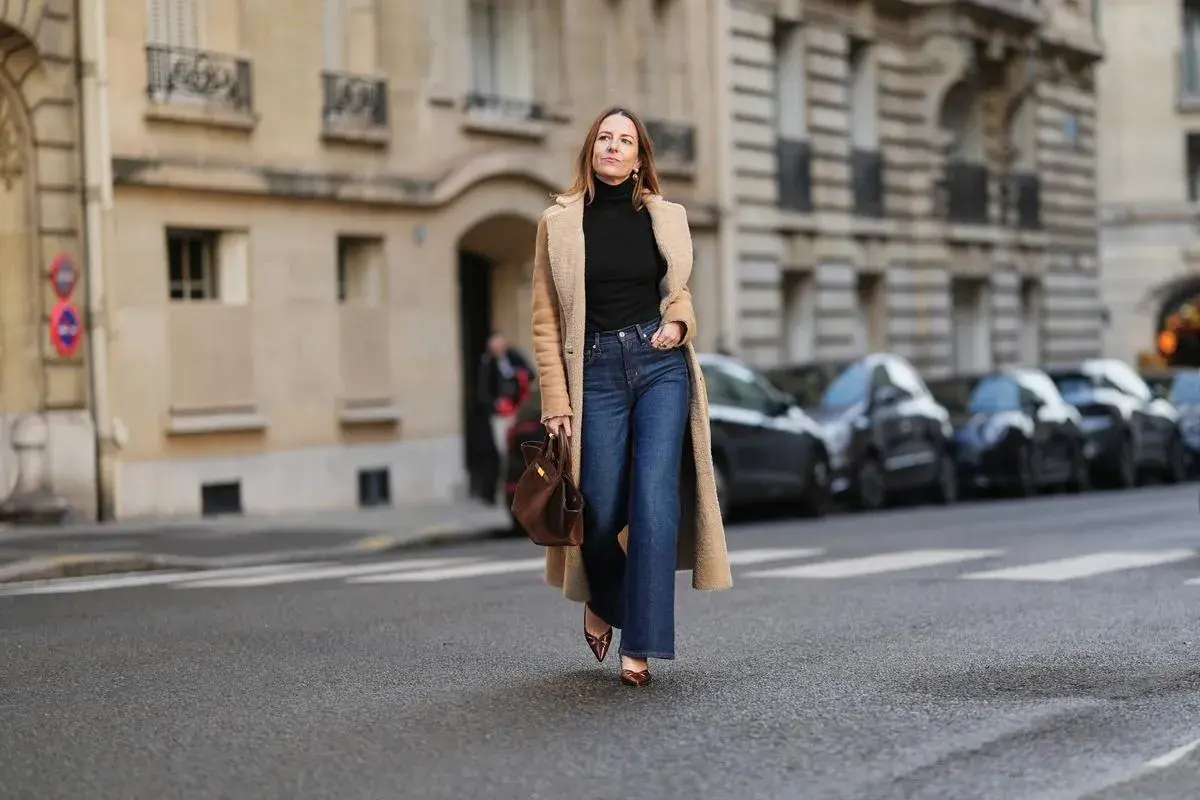 Une femme porte un jean flare bleu, idéal pour allonger la silhouette et sublimer toutes les morphologies. Elle est habillée d'un long manteau camel et d'un col roulé noir.