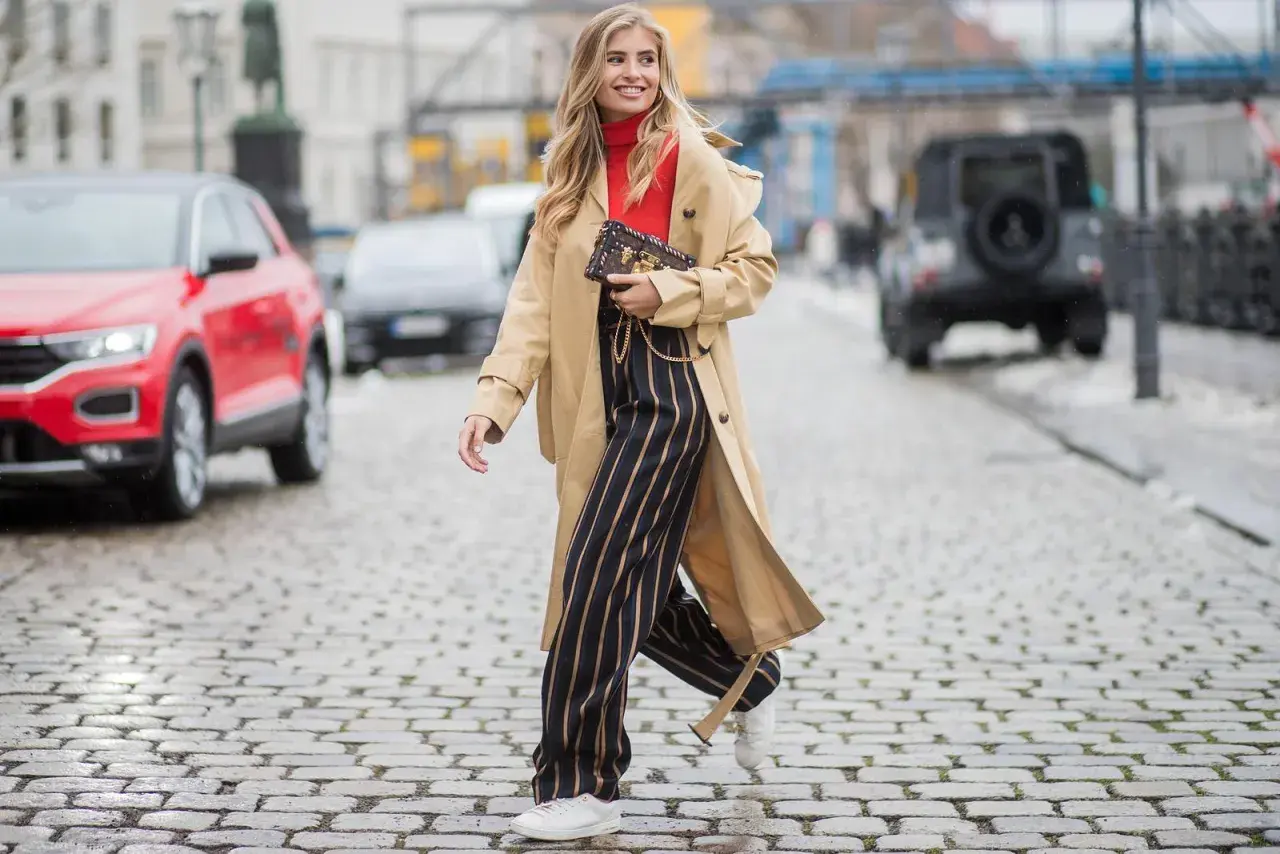 Frau in Beige-Trenchcoat, rotem Rollkragenpullover und gestreiften Hosen, die zeigt, wie man einen Trenchcoat kombinieren kann.