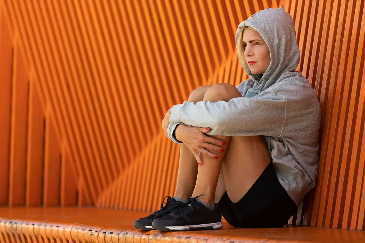 En kvinna i grå hoodie sitter hopkrupen mot en orange vägg, en bild av mental styrka trots en stunds eftertanke.