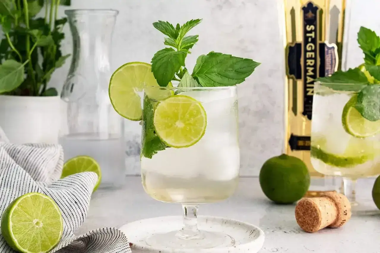 Orzeźwiający Hugo Spritz z limonką i miętą, idealny na letnie popołudnie. Obok butelka St-Germain.
