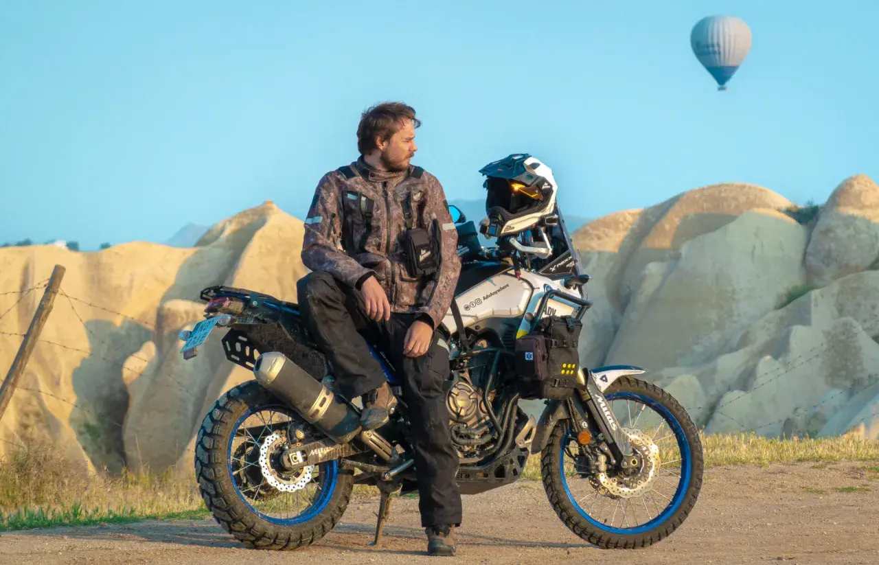 Mężczyzna w kurtce Revit Sand 4 siedzi na motocyklu terenowym na tle skalistych formacji i balonu na gorące powietrze.