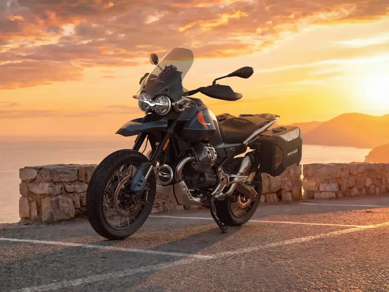Moto Guzzi V85 TT lista para la aventura al atardecer, con su diseño robusto y maletas laterales, frente a un mar sereno y montañas doradas.