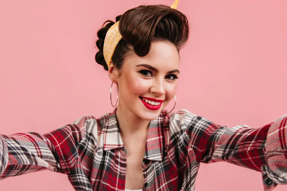 Uśmiechnięta kobieta w stylu pin up, z burzą brązowych włosów upiętych w kok i żółtą apaszką, w kraciastą koszulę i z dużymi kolczykami.