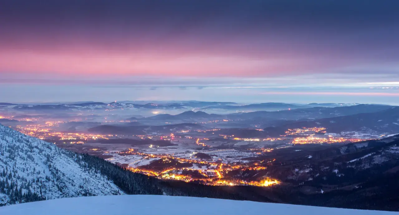 Zachód słońca nad Karkonoszami. Rozświetlone miasto w dolinie, ośnieżone zbocza i zimowe szlaki tworzą magiczny krajobraz.