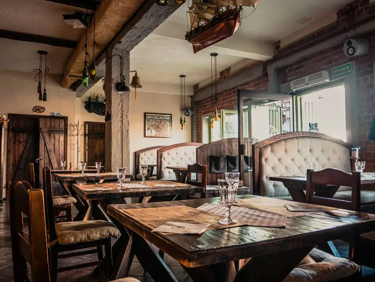 Wnętrze restauracji z drewnianymi stołami i wygodnymi kanapami. Na ścianie wisi model statku, nawiązujący do morskiego klimatu, jak w gdańskim bowke.