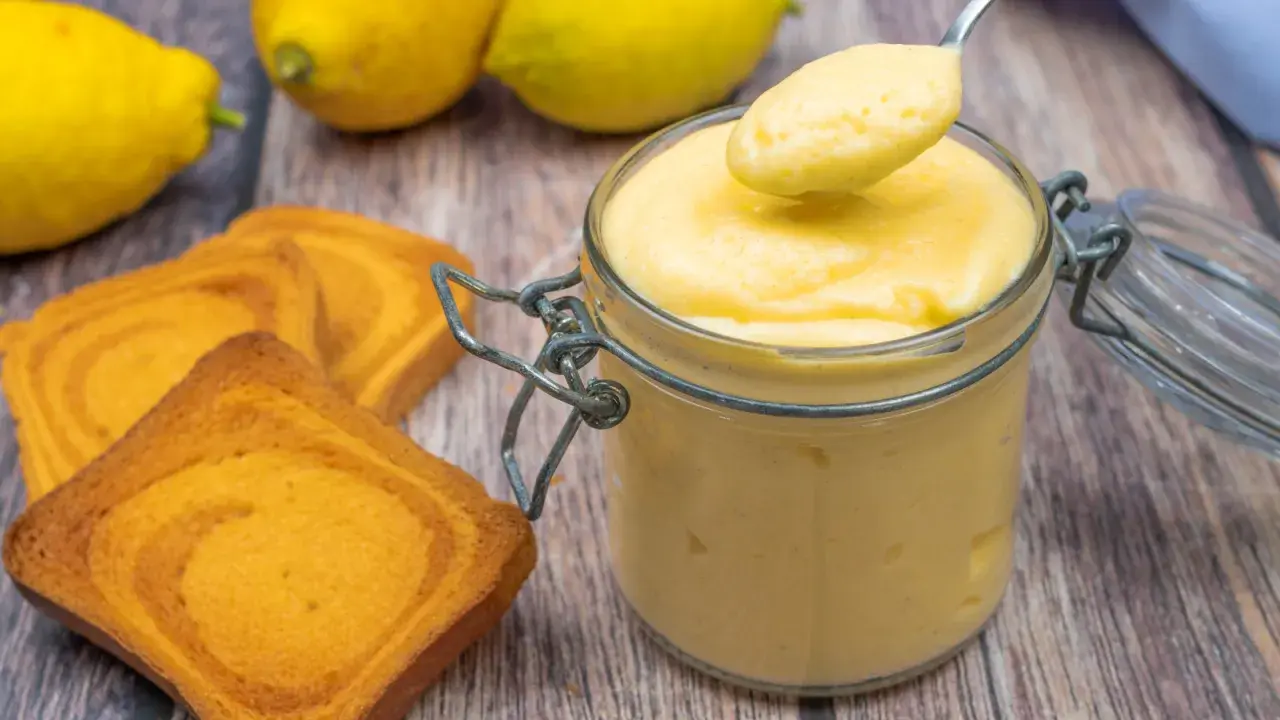 Vaso di vetro pieno di cremoso lemon curd, pronto per essere gustato con biscotti e limoni freschi.