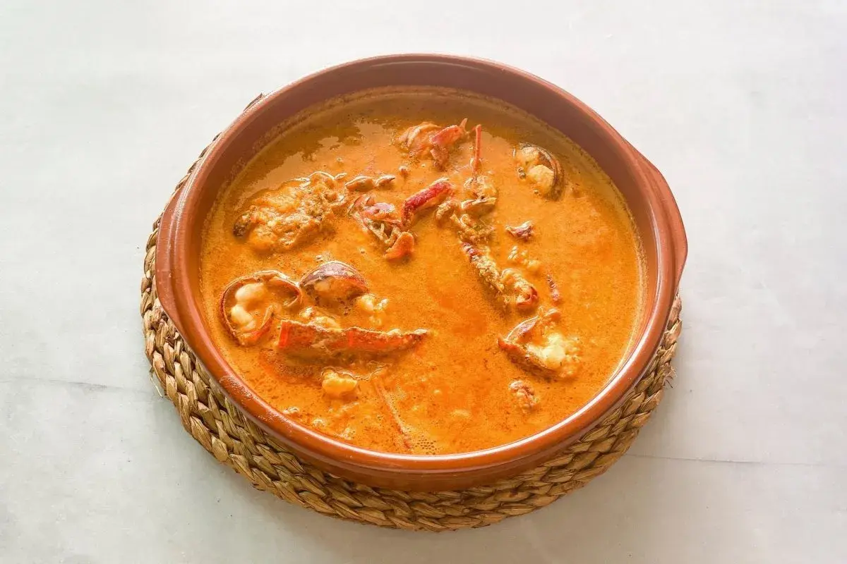 Caldereta de langosta, un plato de marisco reconfortante en una cazuela de barro, con trozos de langosta en una salsa naranja cremosa.