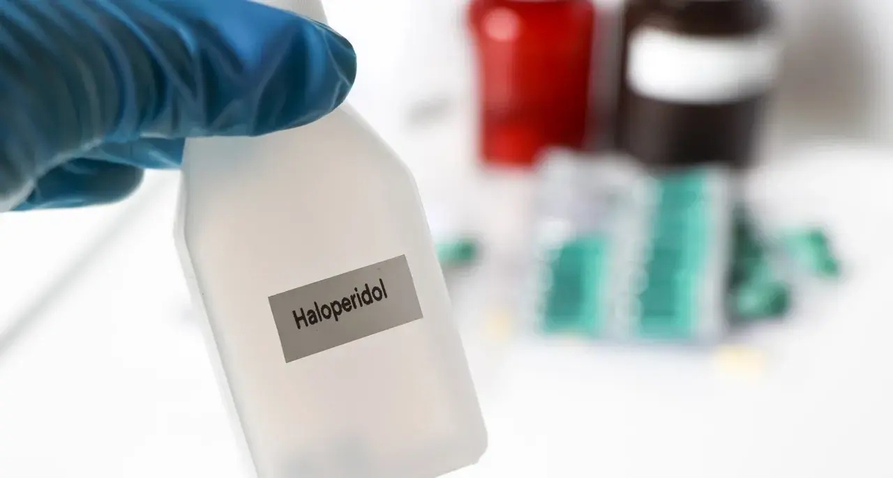 Blauer Handschuh hält Flasche mit der Aufschrift "Haloperidol". Im Hintergrund sind Medikamentenpackungen zu sehen.