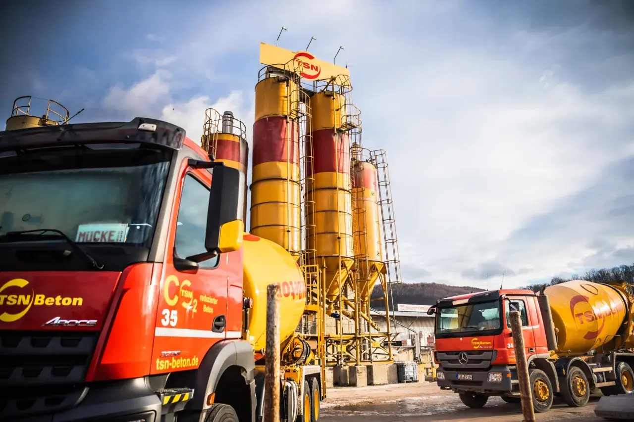 Zwei Betonmischer stehen vor hohen Silos einer Betonmischanlage. Die hgt tragschicht ist bereit für den Transport.