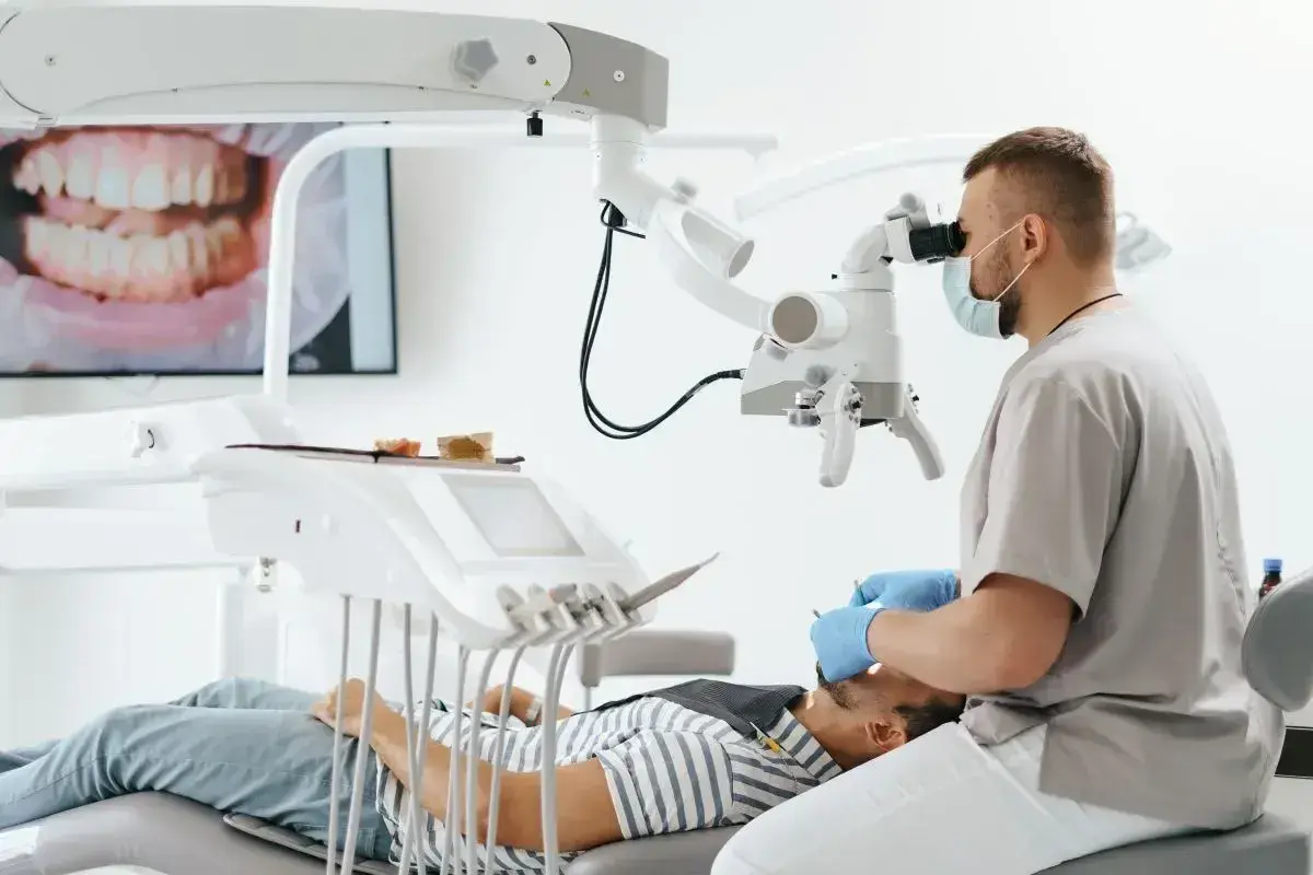Stomatolog w masce i rękawiczkach bada pacjenta pod mikroskopem. Na ekranie widać zbliżenie zębów.