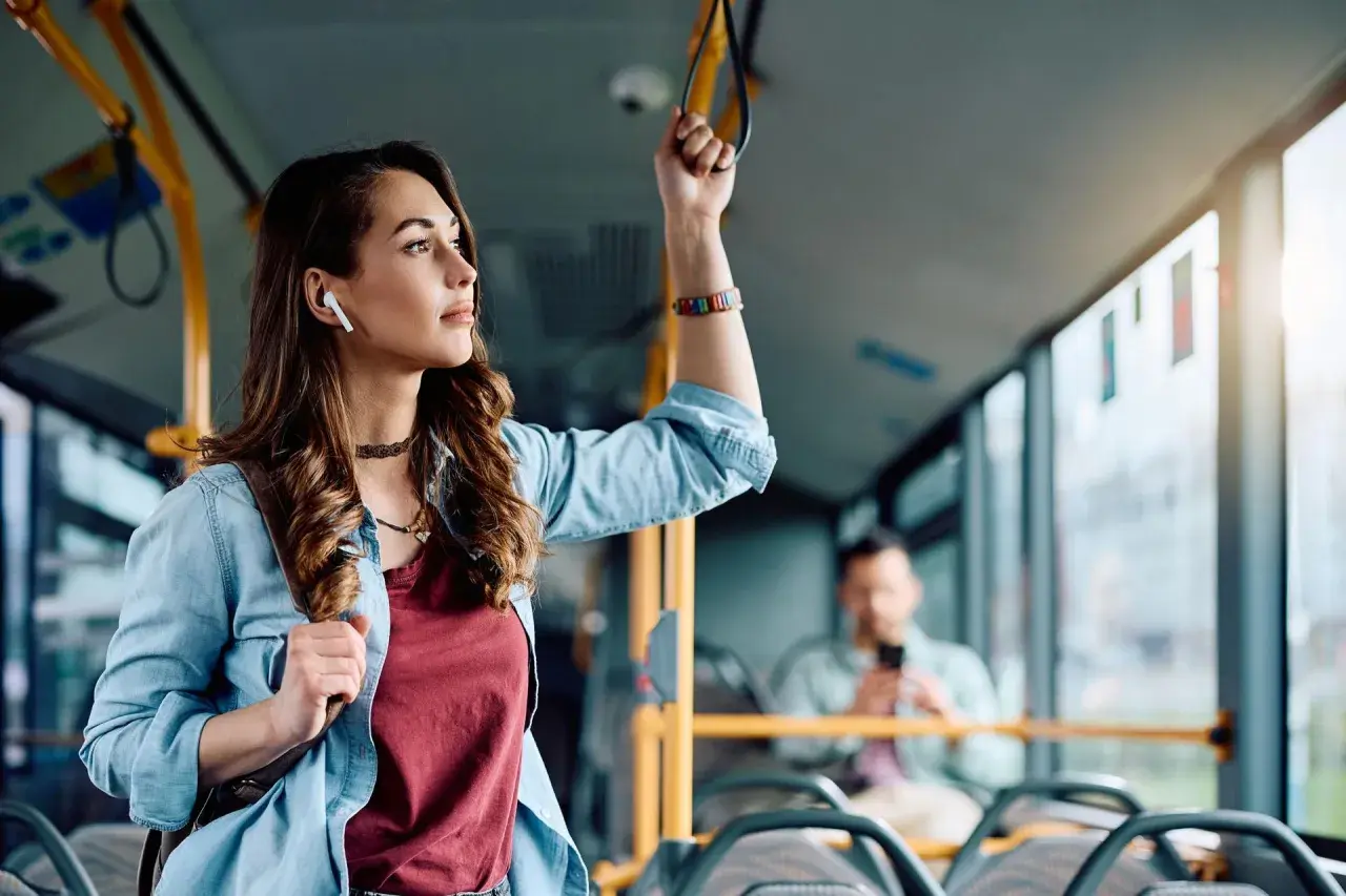 Młoda kobieta w słuchawkach bezprzewodowych trzyma się poręczy w autobusie, słuchając muzyki.