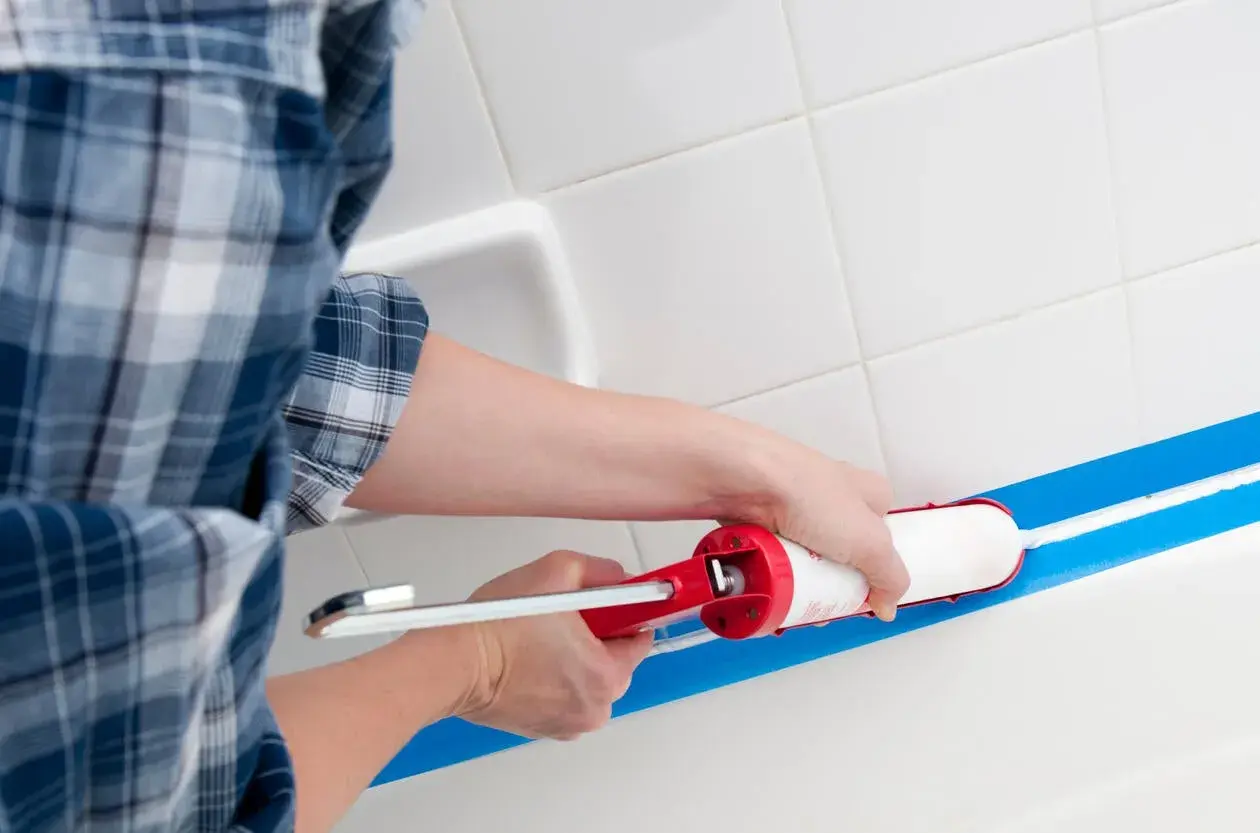 Osoba w kratę zakłada silikon sanitarny w wannie, używając pistoletu i taśmy malarskiej.