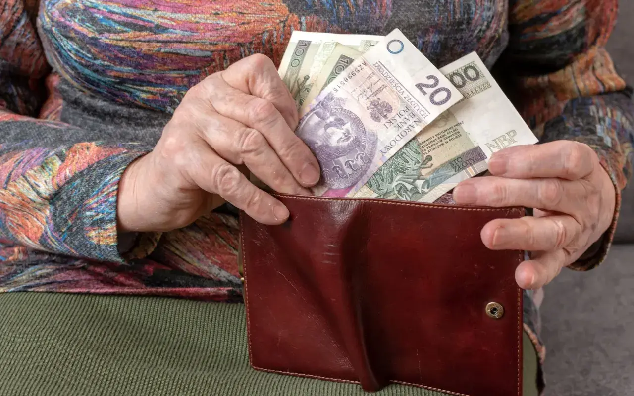 Dłoń starszej osoby wkłada polskie banknoty do brązowego portfela. To pieniądze, ich ekwiwalent.