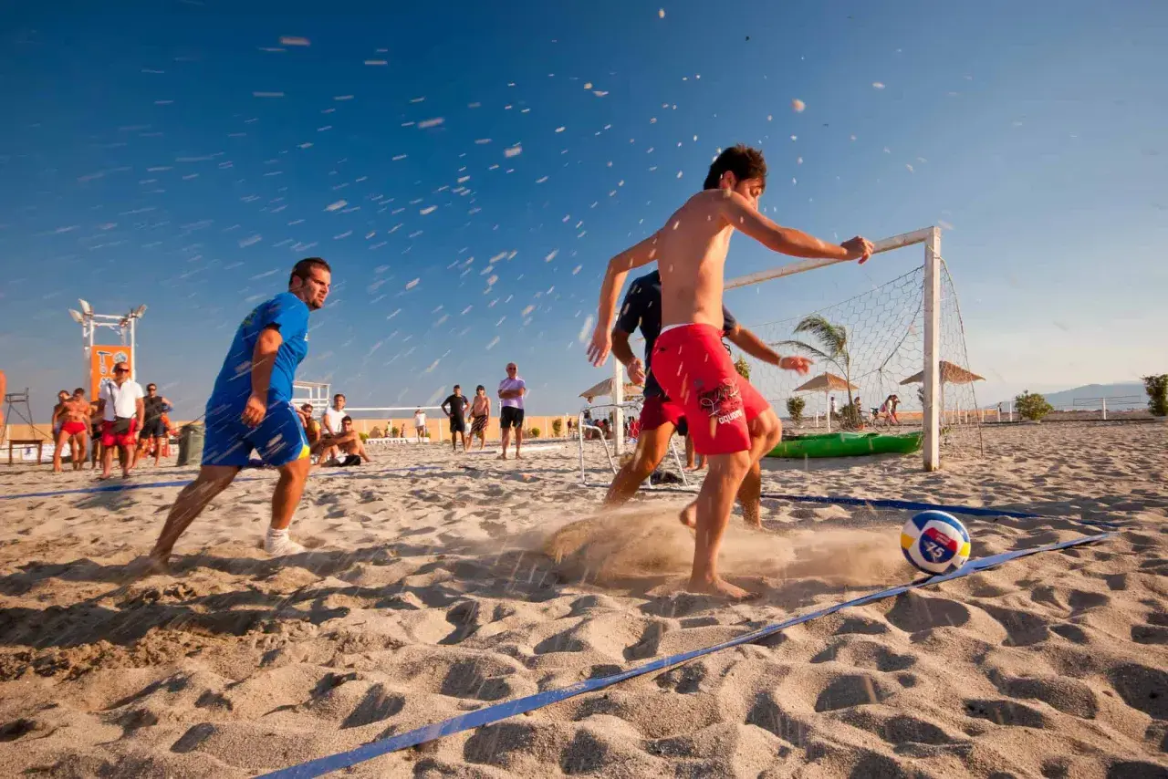 Mężczyźni grają w piłkę nożną na plaży, pokazując różne odmiany tej gry. Słońce, piasek i błękitne niebo tworzą idealne warunki do sportowych emocji.