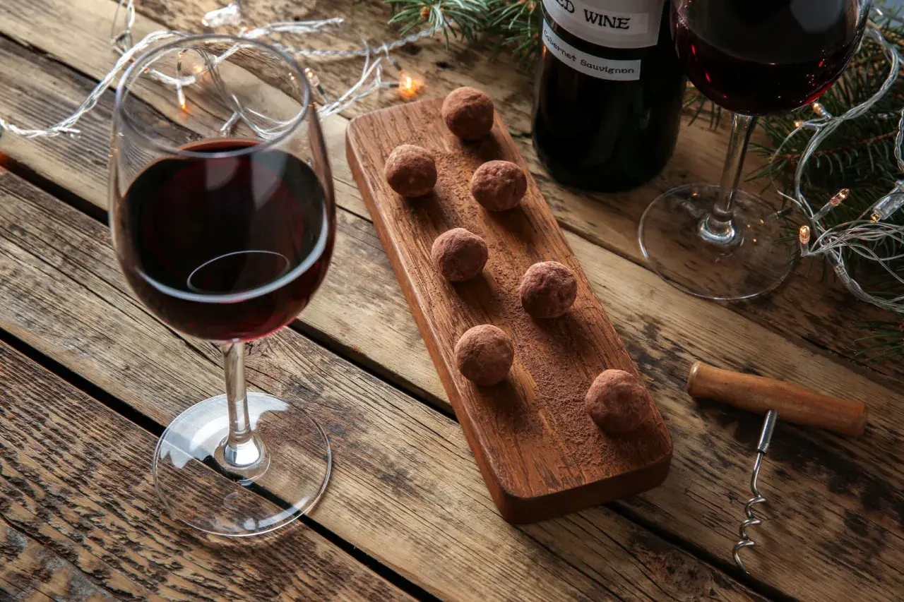 Ein Glas Rotwein und Schokoladentrüffel auf einem Holzbrett, daneben eine Flasche Wein und Lichterketten. Perfekt für einen gemütlichen Abend mit Wein und Schokolade.