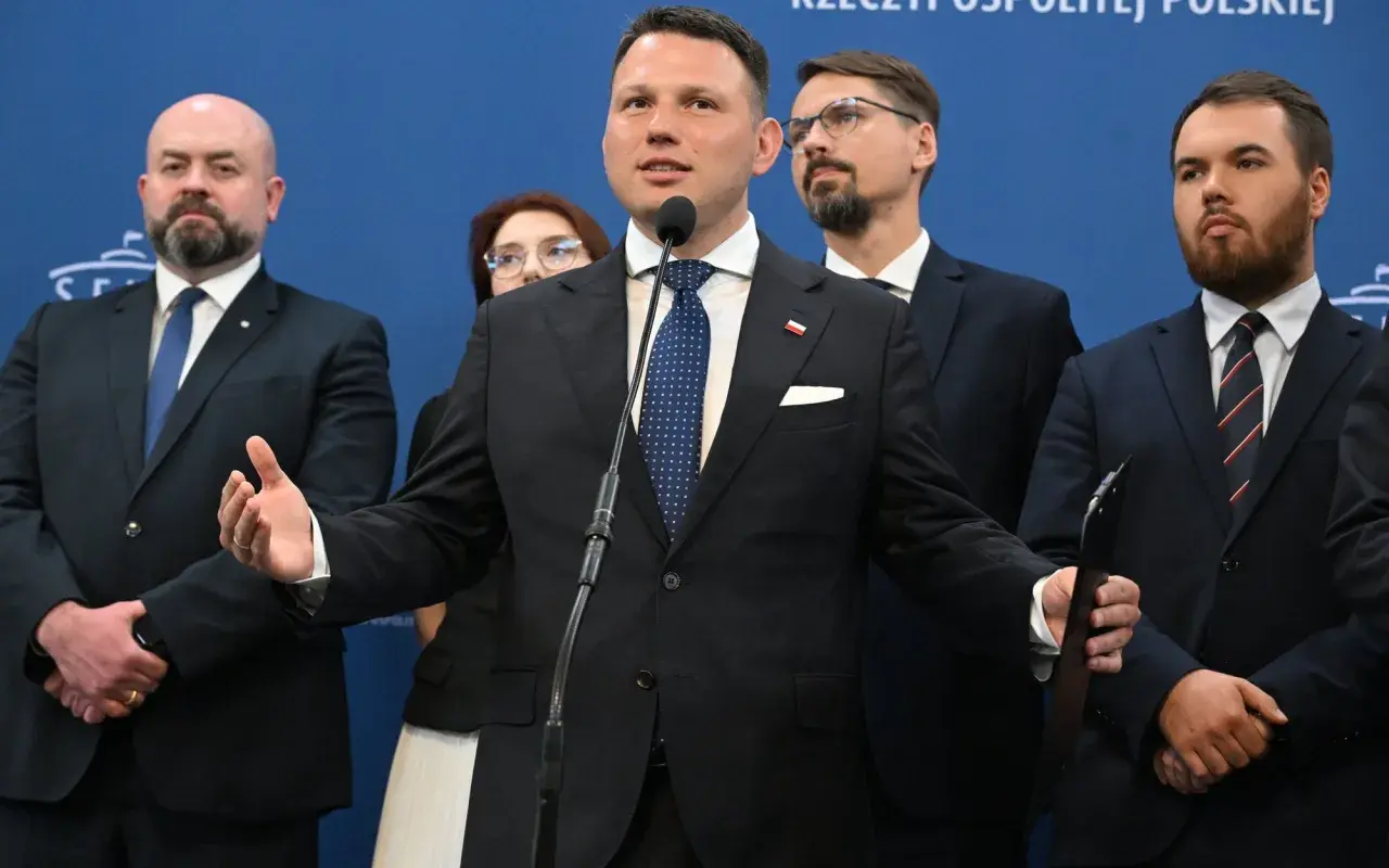 Mężczyzna przemawia przy mikrofonie, otoczony innymi politykami. W tle niebieska ściana z napisem. Konfederacja postulaty.