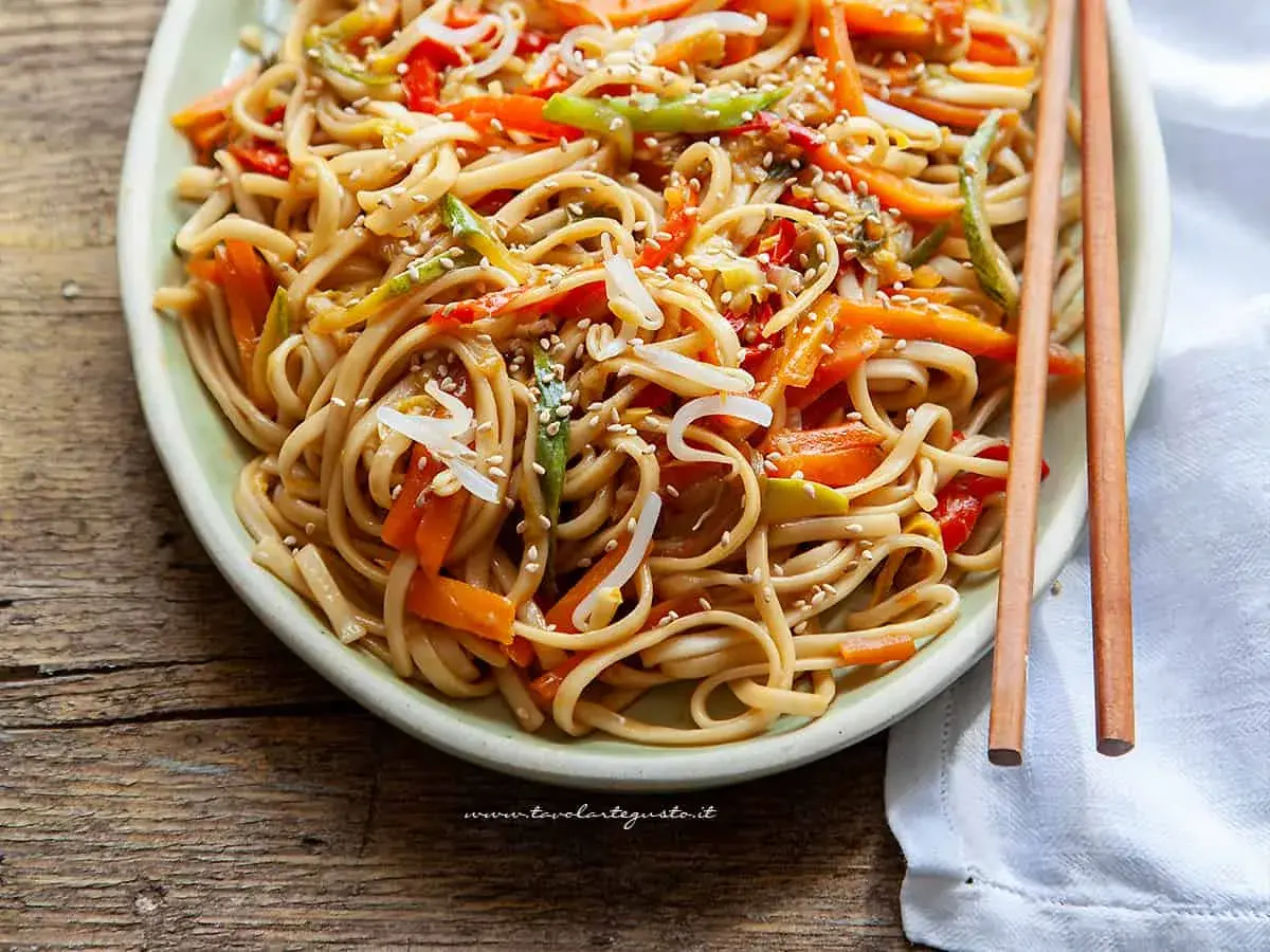 Un piatto invitante di spaghetti di riso saltati con verdure colorate e semi di sesamo, pronti per essere gustati con le bacchette.