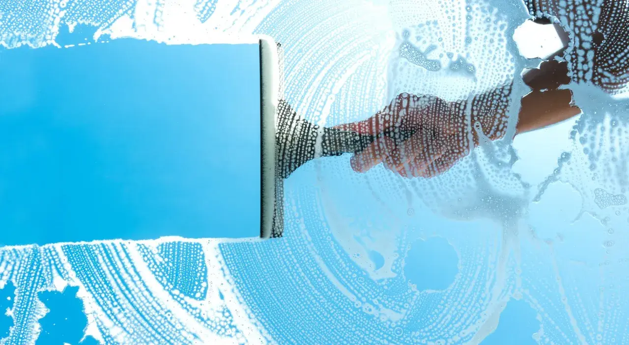 Mano con tergivetro crea strisce di schiuma su un vetro azzurro, per una perfetta pulizia finestre.