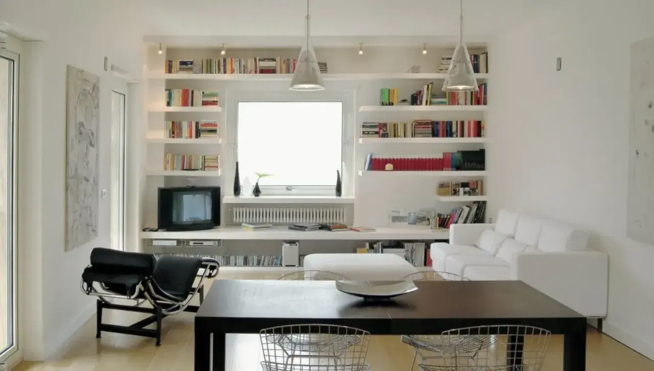 Soggiorno moderno con ampie librerie cartongesso a parete, piene di libri, che incorniciano una finestra. Divano bianco e poltrona nera completano l'arredo.